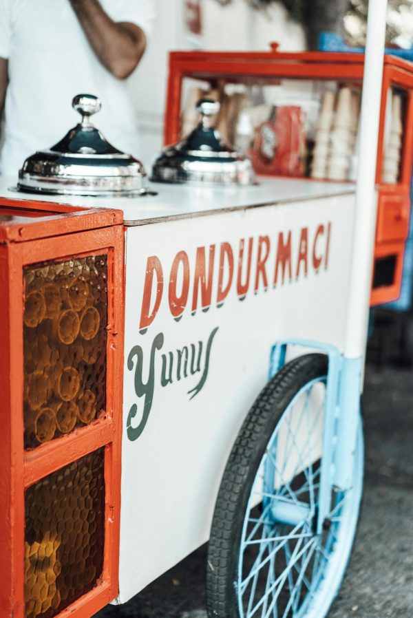 Food cart with metal lids and various ingredients with colorful inscription parked on asphalt sidewalk on street in city on blurred background